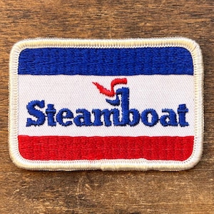 Könnte beinhalten: Rechteckiger, gestickter Aufnäher mit blauem Oberteil, weißer Mitte und rotem Unterteil. Das Wort "Steamboat" ist in blauer Schrift geschrieben. Eine rot-blaue Grafik befindet sich über dem Wort. Weißer Rand.