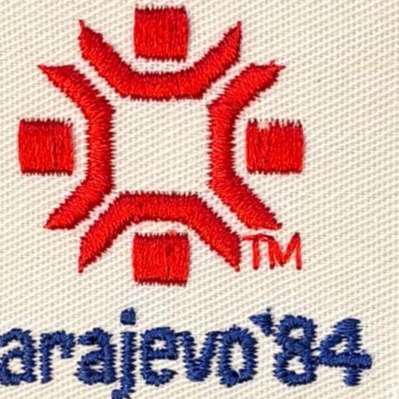 Puede incluir: Parche bordado con un dise&ntilde;o de copo de nieve rojo estilizado y el texto "Sarajevo '84" en azul. El dise&ntilde;o incluye un centro cuadrado y brazos radiantes, con un s&iacute;mbolo "TM".