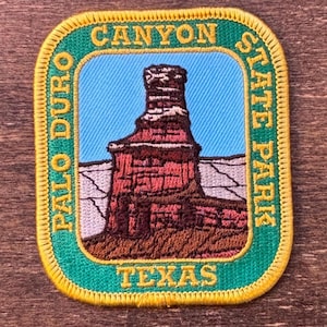 Puede incluir: Parche bordado con un paisaje del Parque Estatal Palo Duro Canyon en Texas. El parche tiene forma rectangular con esquinas redondeadas, un borde verde y las palabras "CANYON STATE PARK" y "PALO DURO TEXAS". La imagen central representa una formación rocosa roja sobre un cielo azul.