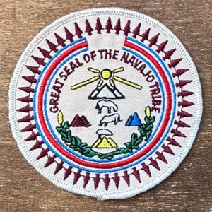 Puede incluir: Parche circular con el Gran Sello de la Tribu Navajo. El diseño incluye un emblema central con montañas, animales y un sol, rodeado de texto y un borde rojo, blanco y azul. El parche está bordeado con formas de flecha granate.