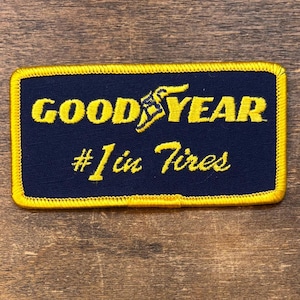 Può includere: Toppa ricamata blu navy con bordo giallo. Presenta le parole "GOOD YEAR" in giallo, con un design ad ala stilizzato, e il testo "#1 in Tires" in corsivo giallo.