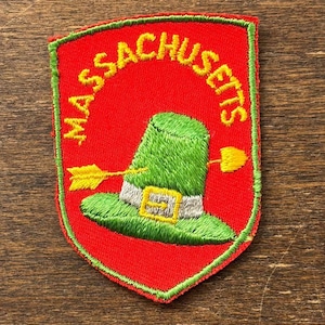 May include: Embroidered patch featuring the word "MASSACHUSETTS" in yellow, arched over a green pilgrim hat pierced by a yellow arrow and shovel. The patch has a red background and a green border.