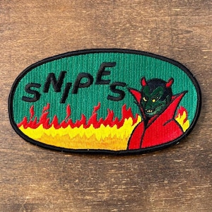 May include: An oval embroidered patch featuring a green background with the black text SNIPES. Below the text are yellow and red flames. On the right side, a green devil figure with horns wears a red cape, all outlined in a thick black border.