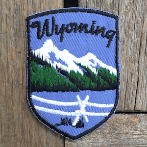 Puede incluir: Un parche bordado azul y blanco con la palabra "Wyoming" en letras negras. El parche presenta una imagen estilizada de una cordillera con picos nevados y árboles verdes.