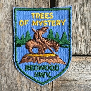 May include: Embroidered patch with a blue background featuring the text "TREES OF MYSTERY" and "REDWOOD HWY." A brown bear-like creature is depicted on a brown mound, with green trees in the background. The patch has a green border.