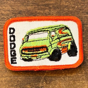 Op de afbeelding: Geborduurde patch met een groene bus met oranje en gele accenten. De bus heeft zwarte wielen en staat tegen een witte achtergrond met een oranje rand. Het woord "DODGE" is verticaal aan de linkerkant geborduurd.
