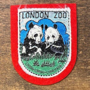 Puede incluir: Un parche bordado en forma de escudo con un borde rojo. El centro muestra dos pandas blancos y negros sentados en hierba verde bajo un cielo azul. El texto LONDON ZOO está bordado en negro en la parte superior, y CHING-CHING-CHIA-CHIA abajo.