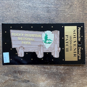 Può includere: Un toppa da stirare ricamata di colore marrone e beige con il testo "Rocky Mountain National Park" e un logo del parco verde e bianco. La toppa è su una carta nera con testo dorato che dice "Pinnacle Designs Souvenir Iron-on Patch".