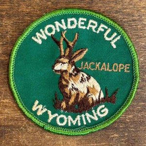 May include: A circular embroidered patch with a green background featuring a brown and white jackalope illustration. The text WONDERFUL is arched at the top, JACKALOPE is in the center, and WYOMING is arched at the bottom in white lettering.