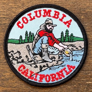 May include: Circular embroidered patch with a black border, depicting a fisherman in a river, with the text "COLUMBIA CALIFORNIA" in red. The design includes trees and a light blue river, set against a light blue background.