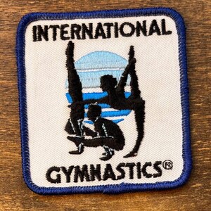 May include: Embroidered patch with the words "INTERNATIONAL GYMNASTICS" in black on a white background. The patch features black silhouettes of gymnasts performing various poses, with a blue and white design in the background. The patch has a blue border.