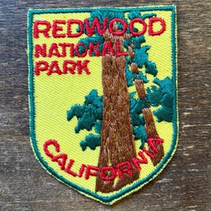 May include: A shield-shaped embroidered patch with a yellow background and a green border. It features a brown redwood tree trunk, dark green foliage, and red embroidered text that reads REDWOOD NATIONAL PARK and CALIFORNIA.