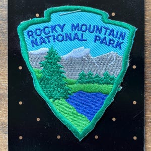 Op de afbeelding: Geborduurde patch met de tekst "Rocky Mountain National Park". De schildvormige patch heeft een turquoise rand en achtergrond. Het ontwerp toont bergen, bomen en een meer, met groen, blauw en grijs.