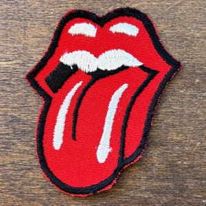 以下が含まれることがあります： ローリング・ストーンズの象徴的な舌と唇のロゴが特徴の赤い刺繍パッチ。パッチは黒い縁取り、白い歯、白い舌があります。このデザインは、ロックンロールの象徴です。