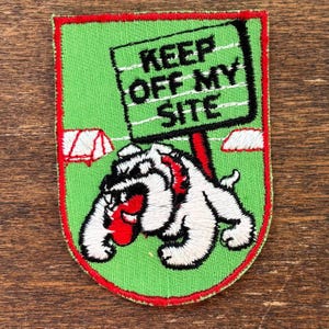 May include: Embroidered patch with a green background and a red border. The patch features a white bulldog holding a sign that reads "KEEP OFF MY SITE". The bulldog has a red collar and is standing in front of a tent.