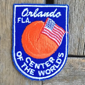Op de afbeelding: Geborduurde patch met een blauwe achtergrond en witte rand. De patch toont de tekst "Orlando FLA." en "Of the Center of the World's" in het wit. Een oranje bol met een rood-wit-blauwe Amerikaanse vlag staat in het midden.