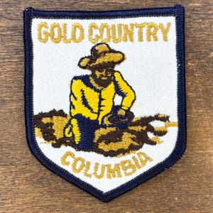 May include: A shield-shaped patch with a white background and a navy blue border. The patch features a gold miner illustration with the text "GOLD COUNTRY COLUMBIA" in gold lettering. The miner is wearing a yellow shirt and hat.