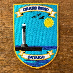 Può includere: Toppa ricamata con un faro, sole e uccelli sull'acqua. La toppa è blu con un bordo giallo e presenta la scritta "GRAND BEND ONTARIO" in bianco.