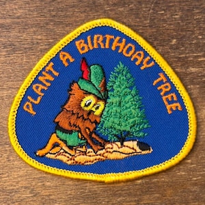 Puede incluir: Parche bordado con fondo azul y borde amarillo. El parche presenta un búho con un sombrero verde plantando un árbol verde. El texto "PLANT A BIRTHDAY TREE" es de color naranja.