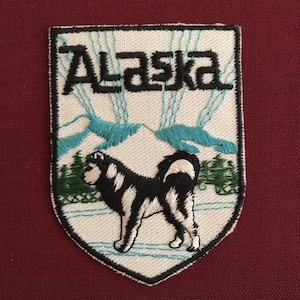 May include: A white embroidered patch with a black and white husky dog standing on a snowy background. The patch features the word "Alaska" in black lettering and a blue mountain range in the background.