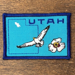 Puede incluir: Parche bordado con el estado de Utah. El parche tiene un borde azul y un fondo turquesa. Muestra la palabra "UTAH", una gaviota volando, un lirio de Sego y una forma blanca que representa el contorno del estado.