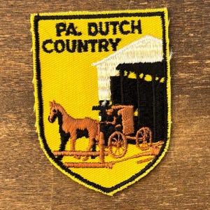 May include: Embroidered patch with a yellow background and black border. The patch features the text "PA. DUTCH COUNTRY" above an image of a horse-drawn carriage and a barn. The horse and carriage are brown and the barn is white.