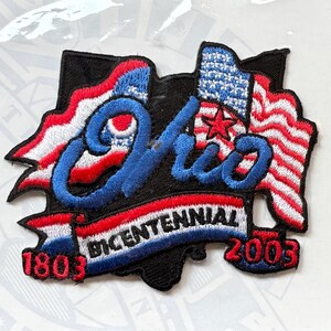May include: Embroidered patch featuring the outline of Ohio in black, with the word "Ohio" in blue script. The patch includes red, white, and blue flags, a red star, and the text "BICENTENNIAL 1803-2003".