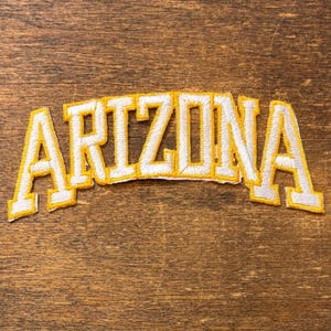 May include: An embroidered patch featuring the word ARIZONA in a bold, arched block font. The letters are white with a thick, vibrant yellow border, creating a high-contrast design suitable for application onto various fabric surfaces.