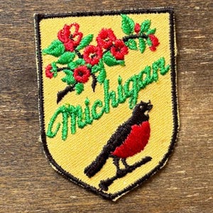 May include: A shield-shaped embroidered patch with a yellow background and a black border. It features red flowers with green leaves at the top, the word Michigan in green cursive script, and a black and red robin bird perched on a branch.