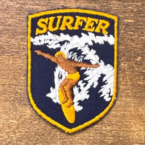 May include: A shield-shaped embroidered patch with a dark blue background and a yellow border. The text SURFER is stitched in yellow at the top. A brown figure is depicted riding a yellow surfboard through white embroidered ocean waves.