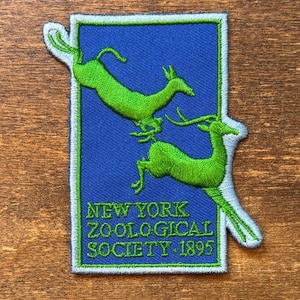Könnte beinhalten: Ein rechteckiger gestickter Aufnäher mit blauem Hintergrund und zwei springenden hellgrünen Hirschen. Der Text NEW YORK ZOOLOGICAL SOCIETY 1895 ist unten in Grün gestickt. Der Aufnäher hat einen hellblauen Rand und unregelmäßige Kanten.