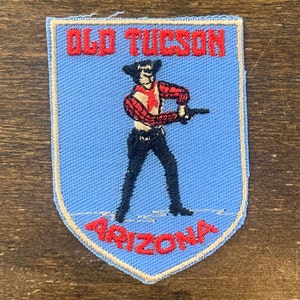 May include: A blue and white embroidered patch with a cowboy holding a gun. The patch says "Old Tucson Arizona" in red lettering.