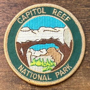 Puede incluir: Parche bordado circular con las palabras "CAPITOL REEF NATIONAL PARK" en un borde verde y dorado. El parche representa un paisaje con cielo azul, formaciones rocosas beige y vegetación verde.