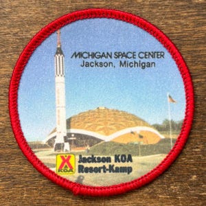 May include: A circular patch with a red border featuring an illustration of the Michigan Space Center in Jackson, Michigan. The image includes a rocket, a domed building, and a small American flag. The text "Michigan Space Center" and "Jackson KOA Resort-Kamp" are also visible.