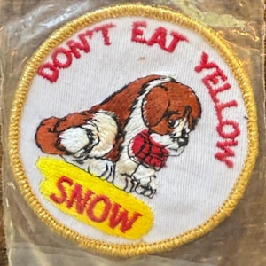 Könnte beinhalten: Gestickter Aufnäher mit einem braun-weißen Cartoon-Hund, der einen roten Schal trägt. Der Aufnäher trägt den Text "DON'T EAT YELLOW SNOW" in Rot und Gelb, mit einem goldenen Rand. Der Aufnäher ist kreisförmig.