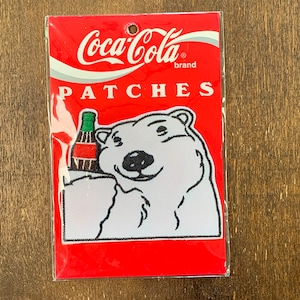 May include: Coca-Cola brand patch featuring a white polar bear holding a bottle of Coca-Cola. The patch is on a red background with white text that says "Coca-Cola brand PATCHES".