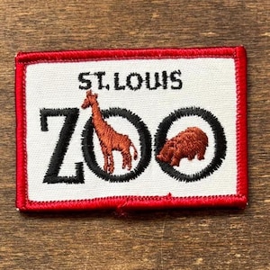 May include: Embroidered patch with a white background and a red border. The text "ST. LOUIS ZOO" is at the top. The word "ZOO" is in black with a giraffe and a bear inside the Os. The giraffe and bear are brown.