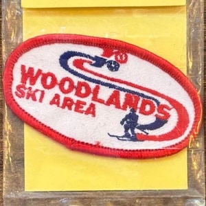 May include: An oval-shaped embroidered patch with a white background and a red border. The text reads WOODLANDS SKI AREA in red letters. A blue graphic depicts a skier in motion, with a red curved line accenting the design of the patch.