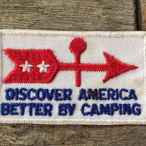 May include: Embroidered patch with a red arrow design and the text "DISCOVER AMERICA BETTER BY CAMPING" in blue. The arrow has two white stars and a red arrowhead. The patch is rectangular with a white background.