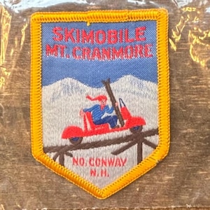May include: Embroidered patch featuring a red skidoo with a person in blue and red clothing. The patch reads "SKIMOBILE MT. CRANMORE" above the skidoo and "NO. CONWAY N.H." below. The patch has a blue background with a snowy mountain scene.