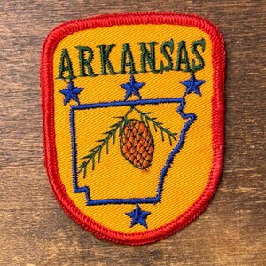 Pode incluir: Um emblema bordado em forma de escudo com fundo amarelo e borda vermelha. Apresenta a palavra ARKANSAS em letras verde-escuras, o contorno azul do estado, três estrelas azuis acima, uma estrela azul abaixo e um gráfico de pinha.