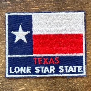 May include: Embroidered patch featuring the Texas state flag. The patch has a blue background with a white star, a white and red striped section, and the words "TEXAS LONE STAR STATE" in white and red.