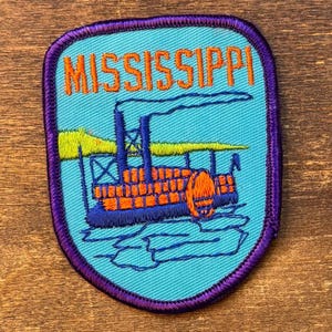 May include: Embroidered patch featuring the word "MISSISSIPPI" in orange above a steamboat illustration. The patch has a teal background with a purple border. The steamboat is orange and blue.