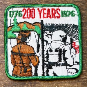 Może przedstawiać: Haftowana naszywka z zieloną obwódką, z napisem "1776 200 YEARS 1976" w kolorze czerwonym i zielonym. Projekt zawiera postać w brązowych ubraniach oraz czarno-białą ilustrację żołnierzy.