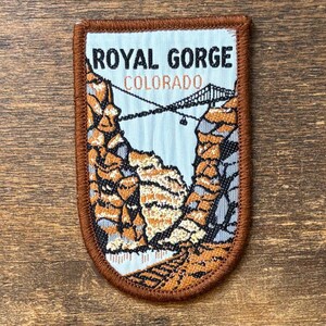 May include: A rectangular embroidered patch with rounded bottom corners featuring a landscape of the Royal Gorge in Colorado. The design includes brown rock formations, a suspension bridge, and the text ROYAL GORGE COLORADO in black and brown.