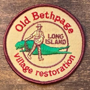Könnte beinhalten: Runder, bestickter Aufnäher mit Text und Bild. Der Text "Old Bethpage Long Island village restoration" ist rot. Das Bild zeigt eine Person, die einen grünen Alligator führt. Der Aufnäher hat einen cremefarbenen Hintergrund und einen weinroten Rand.