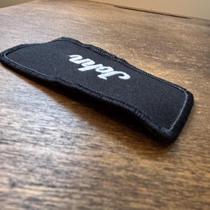 以下が含まれることがあります： 白い筆記体で「John」と刺繍された黒い長方形のパッチ。 パッチは白いステッチの黒い縁取りがあります。 木の表面に置かれたアイテムです。