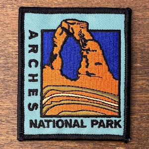 Puede incluir: Parche bordado con el Delicate Arch del Parque Nacional Arches. El parche tiene un borde azul claro con las palabras "ARCHES" y "NATIONAL PARK" en blanco. El arco es naranja sobre un fondo azul.