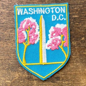 May include: A shield-shaped embroidered patch with a bright blue background and a yellow border. It features white text reading WASHINGTON D.C. above a central white obelisk monument flanked by two stylized pink cherry blossom trees.