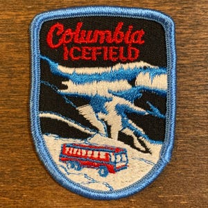 May include: Embroidered patch featuring the words "Columbia ICEFIELD" in red script. The patch depicts a mountain scene with blue and white glaciers, a red bus, and a blue border. The patch is designed to be sewn onto clothing or bags.